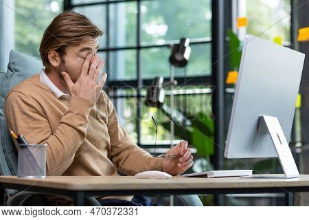 Tired Office Worker Rubs Eyes After Long Work At Computer Inside Office, Young Blond Businessman Ove