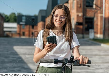 Young Female Riding An Electric Scooter And Holds Smartphone In Hand In Urban Street