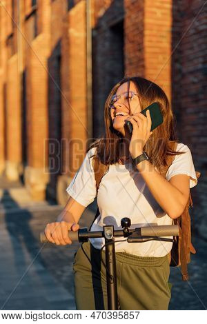Young Female Riding An Electric Scooter And Talking To Smartphone In Urban Street