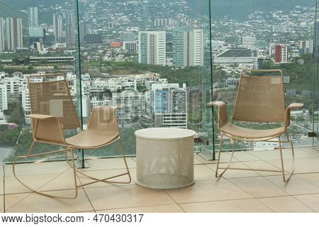 Coffee Table And Rocking Chairs Against Picturesque Landscape Of City In Cafe