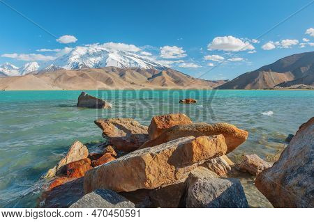 Snowy Peak And A Blue Lake In The Background. Beach In The Rocks. China
