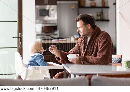 Father Wearing Bathrope Spoon Feeding Hir Infant Baby Boy Child Sitting In High Chair At The Dining 