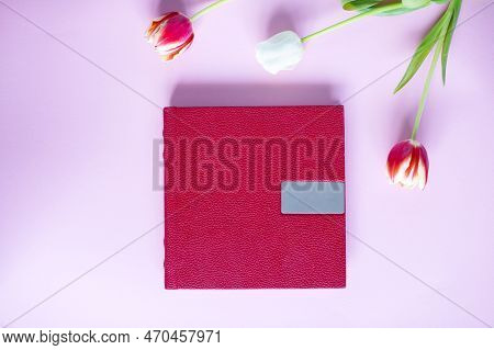 A Red Leather Book With An Embossed Place For Text, Lies On A Purple Background Next To Flowers And 