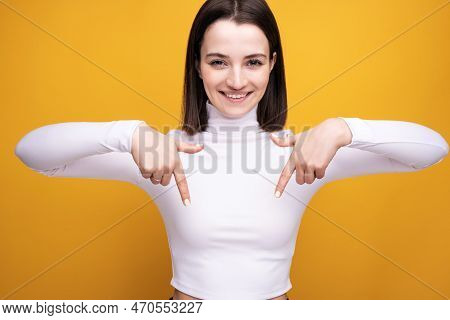 The Woman Smiles And Points Down. Portrait Of A Girl In A White Blouse On A Yellow Background In The