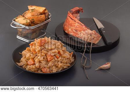 Stewed Cabbage - Bigos In A Black Plate On The Background Of A Piece Of Smoked Meat And Baked Potato
