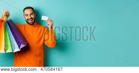 Happy Aduly Man Showing Credit Card And Shopping Bags, Standing Against Turquoise Background