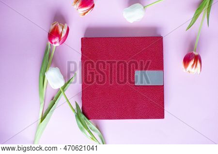 A Red Leather Book With An Embossed Place For Text, Lies On A Purple Background Next To Flowers And 