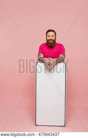 Bearded And Tattooed Man In Magenta T-shirt Looking At Camera Near Huge Phone Template On Pink Backg