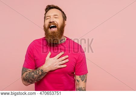 Joyful Bearded Man In Bright T-shirt Touching Chest And Laughing With Closed Eyes Isolated On Pink.