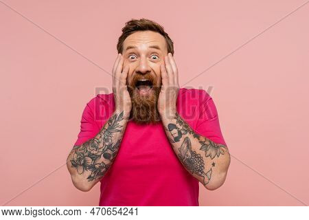 Amazed Bearded Man In Bright T-shirt Touching Face While Looking At Camera Isolated On Pink.