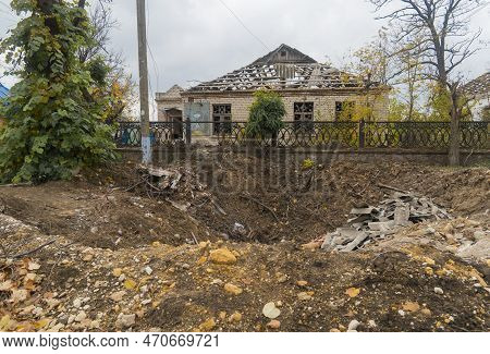Countryside. Crater In The Ground From A Bomb Explosion Against The Background Of A House Destroyed 