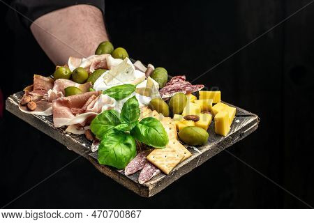 Hands Holding A Glass Of Wine And A Wooden Board With Different Kinds Of Cheese And Ham, Prosciutto,