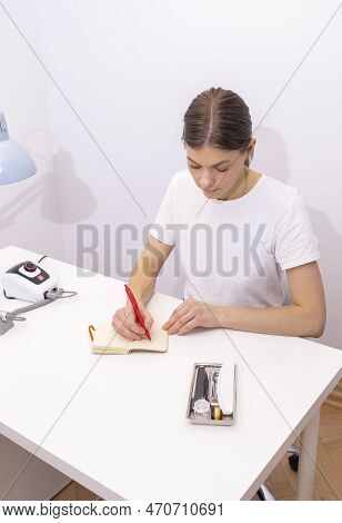 Nail Technician Makes Notes In Notebook With Pen, Sitting At Workstation, Table In Nail Beauty Treat