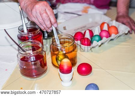 Old Woman Dyeing Easter Eggs At Home In The Kitchen In The Cucumber Glass And With Different Colors,