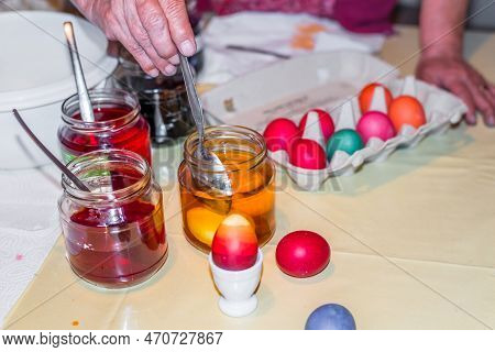 Old Woman Dyeing Easter Eggs At Home In The Kitchen In The Cucumber Glass And With Different Colors,