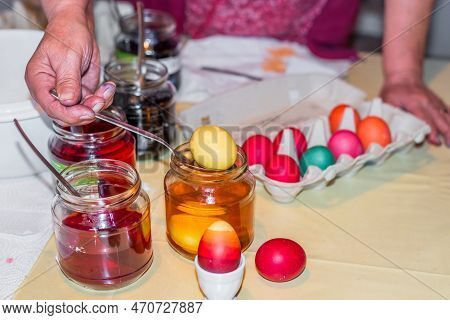 Old Woman Dyeing Easter Eggs At Home In The Kitchen In The Cucumber Glass And With Different Colors,