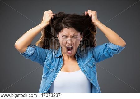An Emotionally Angry Woman In Casual Clothes Screams And Pulls Her Hair Out In Front Of A Gray Wall.