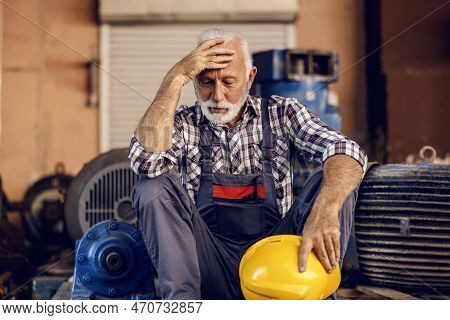 A Sad Senior Factory Worker Sits Next To The Machines With A Helmet In His Hands And Holding His Hea
