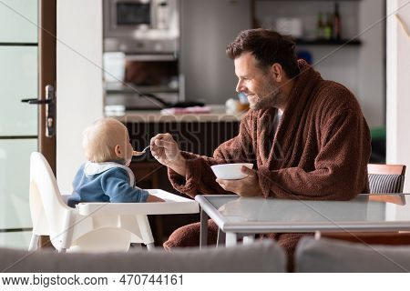 Father Wearing Bathrope Spoon Feeding Hir Infant Baby Boy Child Sitting In High Chair At The Dining 
