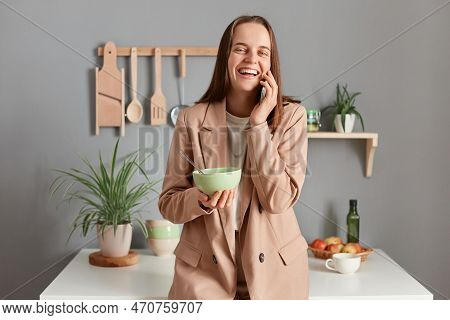 Image Of Attractive Laughing Brown Haired Woman Wearing Beige Suit Standing Near Table On Kitchen At