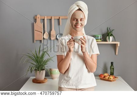 Indoor Shot Of Young Relaxed Woman With Cosmetic Mask In Bath Towel On Her Head Drinking Hot Beverag