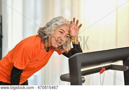 Tired Senior Woman Wiping Sweat Of Her Forehead After Rinning On A Treadmill Indoors. Healthy Active