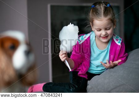 Portrait Of Smiling Little Girl Holding Candyfloss, Looking At Sticky Fingers Hand, Sitting On Sofa 