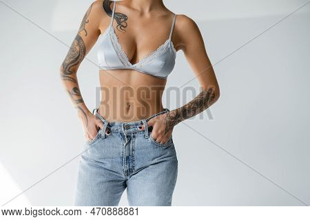 Partial View Of Tattooed And Slim Woman In Silk Bralette Holding Hands In Pockets Of Blue Jeans On G