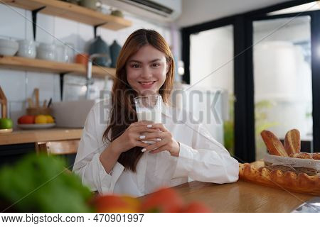 Portrait Of A Young Cheerful Asian Woman Drinking Milk In Kitchen Room With Healthy Raw Food. Vegeta