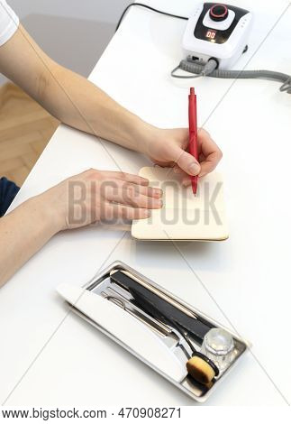 Left Handed Nail Master Makes Notes In Notebook With Pen, Sitting At Workstation, Table In Nail Beau