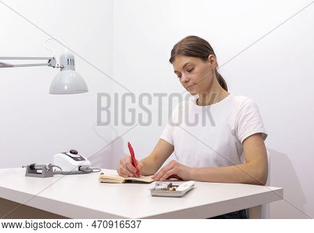 Beautiful Nail Beautician Makes Notes In Notebook With Pen, Sitting At Workstation, Table In Nail Be