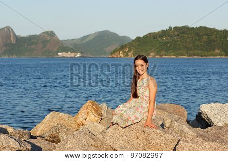 Happy Smiling Beautiful Teen Girl In Dress With Long Brown Hair Sitting On Stones On Beach