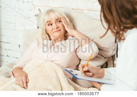 Suffering from horrible pain. Puzzled disappointed aged woman sitting on the bed in the hospital and suffering from headache while listening to the doctors recommendations