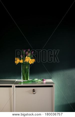 yellow daffodil flowers with pupple tulip blooming in vase with green wall. Dark key. Interior with candle