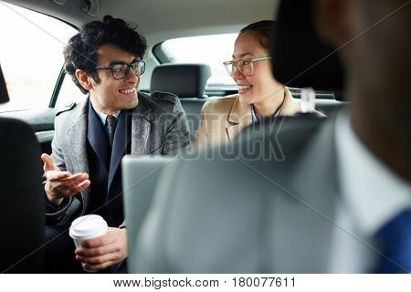Portrait of two successful business people riding on backseat of taxi: smiling cheerfully and talking on the way to work