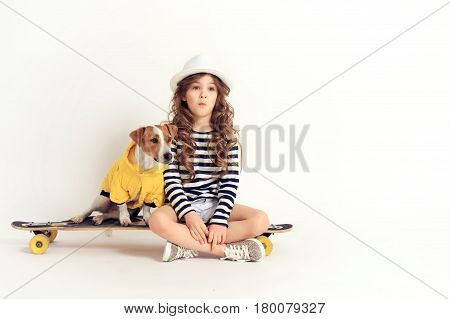 Surprised young girl is sitting on a skateboard with a dog Jack Russell Terrier breed. Picture is taken at the studio with a white colored background.