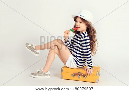 Pretty well-dressed girl is sitting on a yellow suitcase with crossed legs and licking a watermelon lollipop. Picture is taken at studio and has white background. Summer vacation, travel, tourism concept