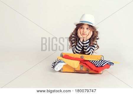 Pretty girl is laying on her filled with coloful clothes suitcase and holding her chin. The picture is taken at studio and has white background.