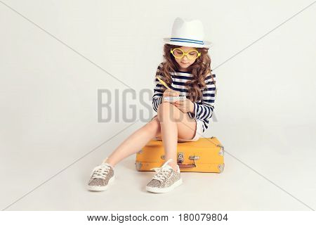 Pretty girl in yellow glasses is writing something in her notebook. She is sitting on a suitcase. The picture is taken at studio and has white background.