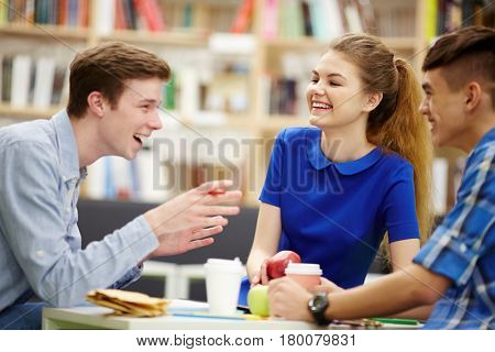 Group of college students talking  in library hall at lunch, sitting round table and laughing, gesturing actively