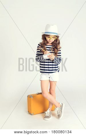 A serious young girl is putting down some marks in her notebook and standing near her yellow suitcase. The picture is taken at studio and has white background.