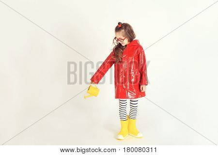A young girl wearing raincoat and rubber boots is pouring water from watering can on invisible plant. The picture is taken at studio and has white background. Childhood, fashion, imagination concept