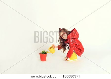 A sitting little girl in red raincoat and rubber boots is watering grass from a pot by using watering can. The picture is taken at studio and has white background