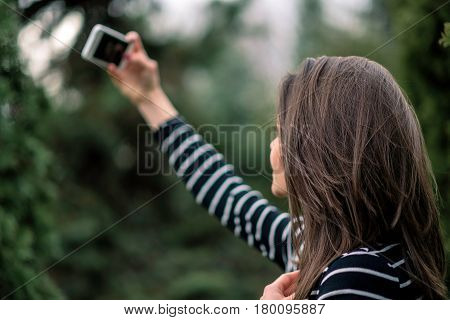 Young girl in striped sweater is taking picture of herself in the park. Shallow depth of field.