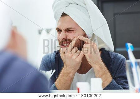Young man sqeezing pimple in front of mirror