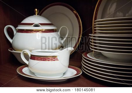 Closeup of white decorated china teaset and piled plates on wooden shelf