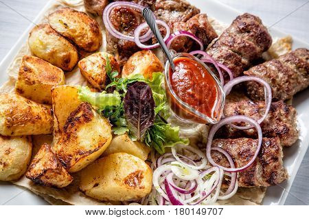 Lyulya kebab with red sauce onion and potatoes on white plate close up with selective focus.