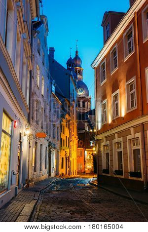 Riga, Latvia. View Of Deserted Narrow Cobbled Kramu Street Of Old Town In Bright Evening Illumination With Ancient Architecture Leading To Dome Cathedral Under Summer Blue Sky.