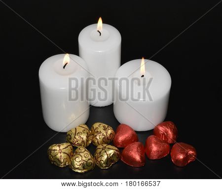 On a black background there are three burning white candles and chocolate sweets in golden and red wrappers