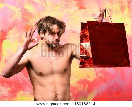 Bearded man short beard. Caucasian sexy macho man with moustache shows six packs and abs on muscle torso holding red package or bag on colorful studio background